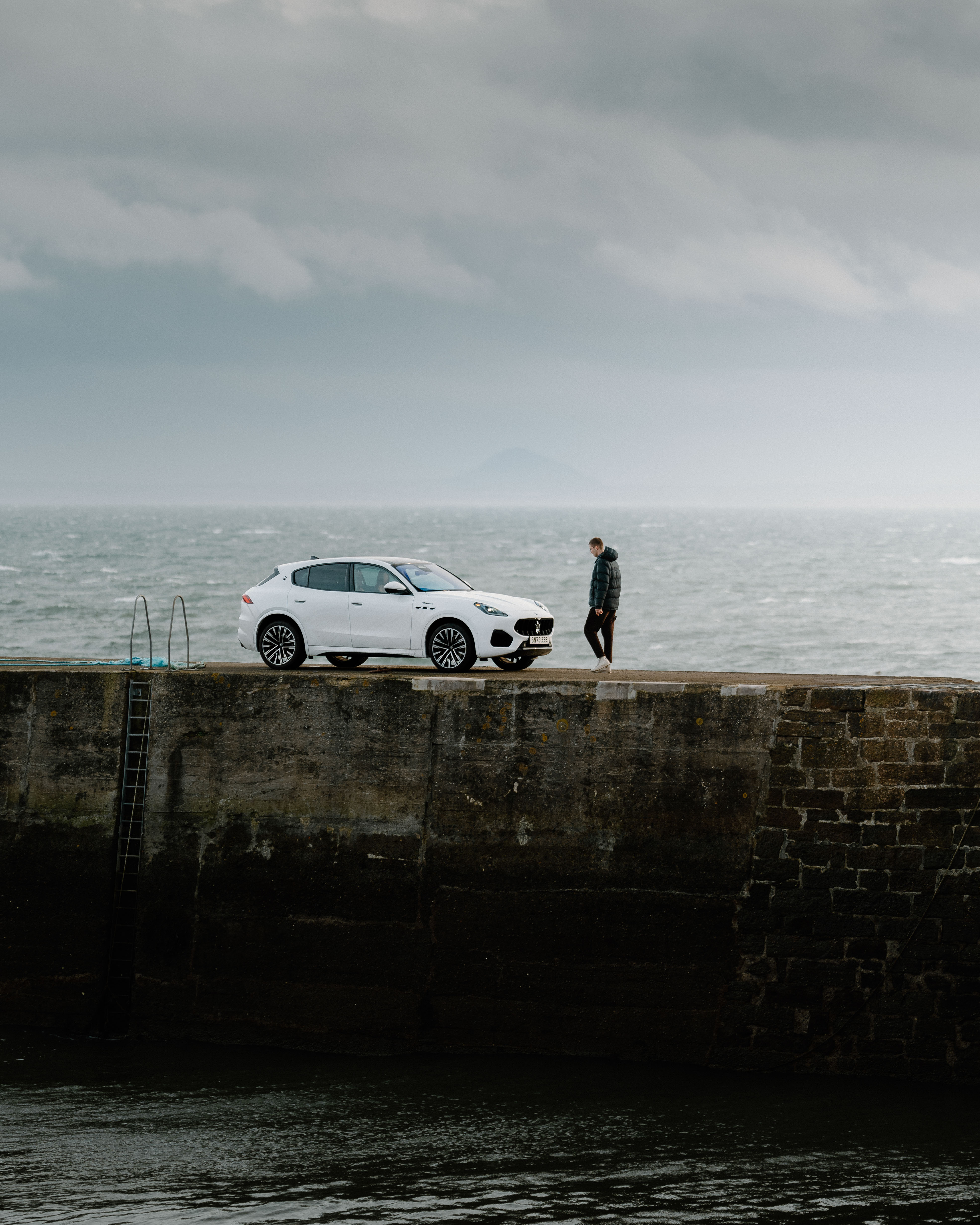 A man stands facing a white car on a pier overlooking the ocean
